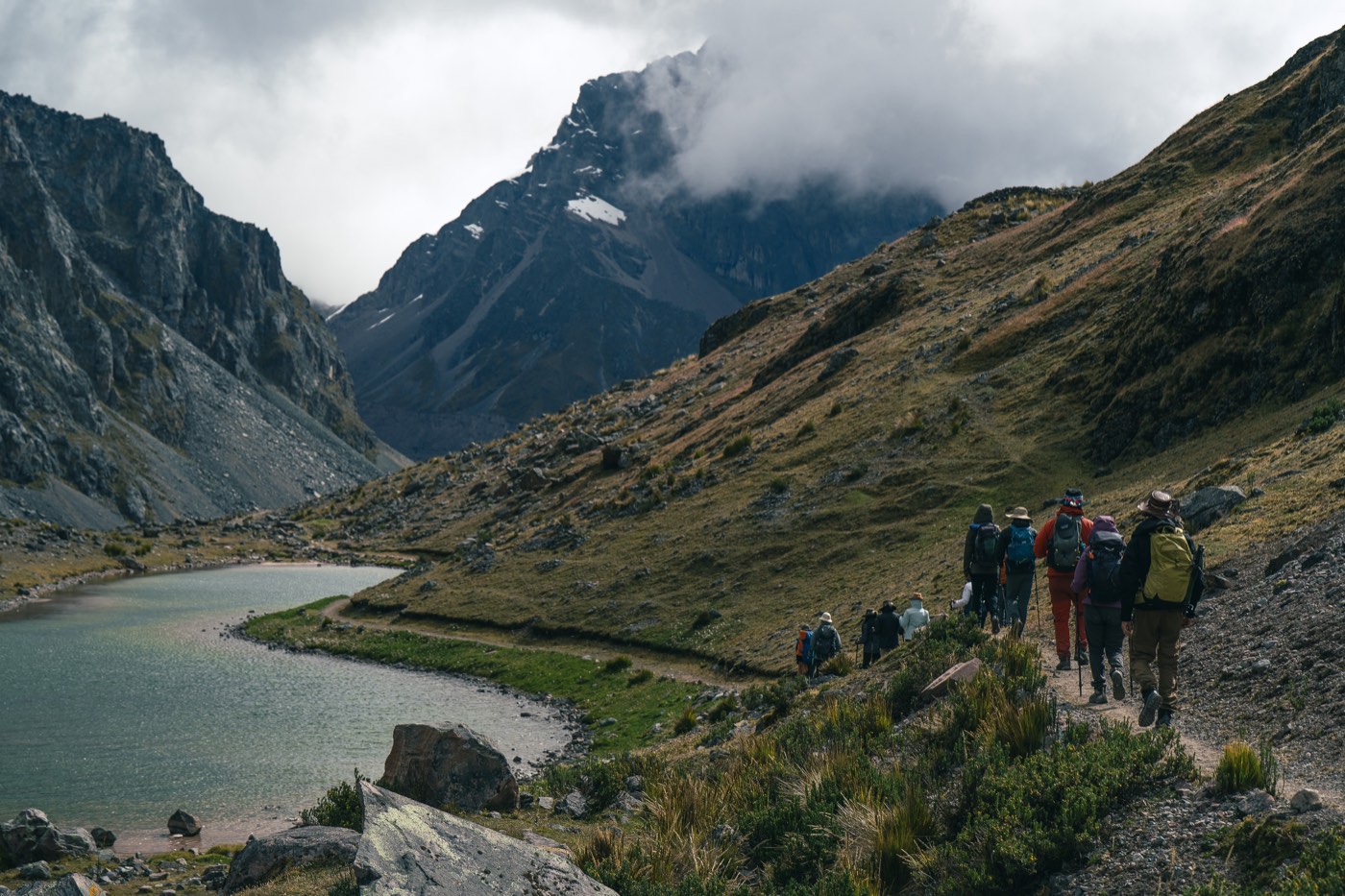 Peregrinos descienden por un sendero junto a una laguna de montaña.